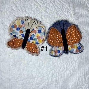 May include: Two fabric butterfly appliques with frayed edges and black central bodies. The left butterfly features multicolored polka dots and solid orange wings. The right butterfly has orange wings with white polka dots and multicolored wing tips. Text #1.