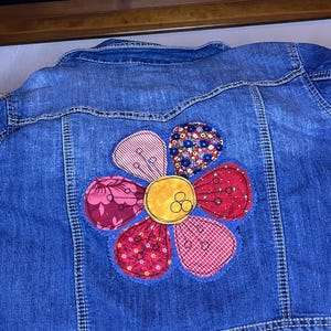 May include: Blue denim jacket with a large, colorful flower applique on the back. The flower features petals in various patterned fabrics, including stripes, floral prints, and gingham, with a yellow center.