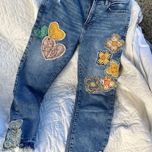 May include: Blue denim jeans with decorative patches. The patches include heart and flower designs in various colors and patterns. The jeans have a distressed look with a few small tears.