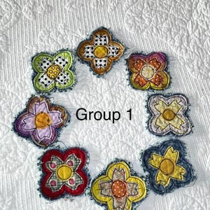 May include: Seven fabric flower appliques arranged in a circle, each featuring four petals with frayed edges and a central circular core. The flowers display various colors including green, yellow, purple, red, and blue with patterned fabric details. The text Group 1 is centered.