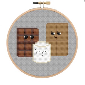 May include: A cross-stitch pattern featuring a chocolate bar, marshmallow, and graham cracker, each with a cute cartoon face. The pattern is displayed in a wooden embroidery hoop.