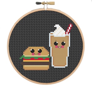 Puede incluir: Un aro de bordado de punto de cruz con un fondo negro que presenta una hamburguesa y un batido de dibujos animados. La hamburguesa tiene un pan marrón, una hoja de lechuga verde, un tomate rojo y una rebanada de queso amarillo. El batido es marrón con una cobertura de crema batida blanca y una pajita roja.