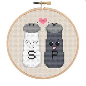 Puede incluir: Un aro de bordado de punto de cruz con un diseño de salero y pimentero. Los saleros están representados como personajes de dibujos animados lindos con caras y las letras "S" y "P" en ellos. Un corazón rosa está por encima de los saleros.