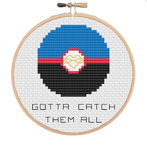 May include: A cross-stitch embroidery hoop featuring a pixelated design in blue, red, and black, with a gold heart-shaped emblem. The text "GOTTA CATCH THEM ALL" is stitched below the design, set against a white background.