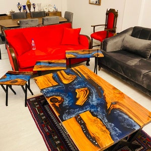 Puede incluir: Una mesa de centro de madera grande con una incrustación de resina epoxi azul y blanca. La mesa tiene una base de metal negro y está sentada sobre una alfombra con un patrón colorido. Hay tres mesas más pequeñas con incrustaciones de resina epoxi similares.