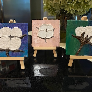 May include: Three small canvas paintings of cotton blossoms on easels. The paintings are in shades of blue, pink, and green. Each painting has the artist's initials "CCR" in the bottom right corner.