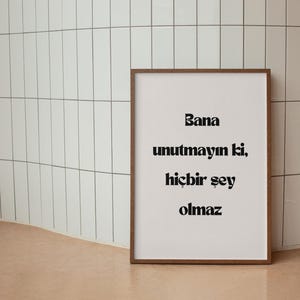 May include: A framed print with the words "Bana unutmayın ki, hiçbir şey olmaz" in black font. The frame is brown, and the background is white. The print is leaning against a white tiled wall.