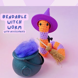 May include: A crocheted witch worm wearing a purple hat and holding a broom. The worm is orange and purple and is sitting on a pink surface. There is a cauldron with purple yarn in it.
