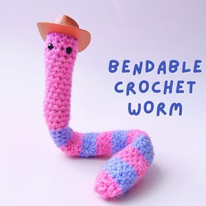 May include: A pink and purple crocheted worm wearing a brown cowboy hat. The worm is bendable and has black eyes. The text "Bendable Crochet Worm" is written in blue letters on a white background.