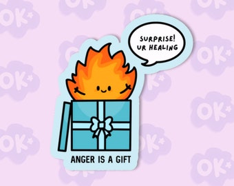 May include: A blue gift box with a white ribbon and a smiling orange fire character inside. The box has the text "ANGER IS A GIFT" on the bottom. A speech bubble above the box says "SURPRISE! UR HEALING".