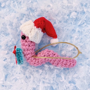 May include: A pink crocheted worm wearing a red and white Santa hat and holding a small green Christmas tree. The worm is on a white background.