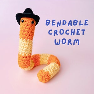 May include: A crocheted orange and yellow worm with a black cowboy hat. The worm is bendable and has googly eyes. The text "Bendable Crochet Worm" is written in blue letters on a pink background.