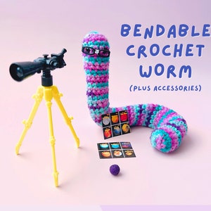 May include: A crocheted worm with a purple, blue, and turquoise striped body is wearing glasses and holding a set of planet cards. The worm is sitting next to a yellow tripod with a black telescope.
