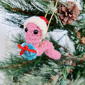 May include: Pink crocheted worm wearing a white crocheted Santa hat and holding a blue and white wreath with a red bow. The worm is hanging from a gold string on a green Christmas tree.