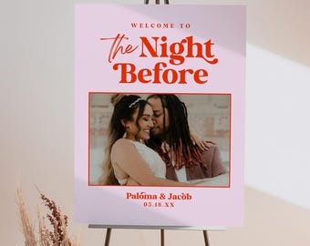 May include: A pink wedding welcome sign on a black easel. The sign reads "WELCOME TO The Night Before" in red script. Below is a photo of a couple, with the names "Paloma & Jacob" and the date.