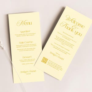 Peut inclure: Deux cartons de menu de mariage crème avec du texte doré. L'une des cartes indique "Menu" avec une liste de plats. L'autre carte dit "Bienvenue et Merci". Une brindille d'herbe séchée est à gauche.