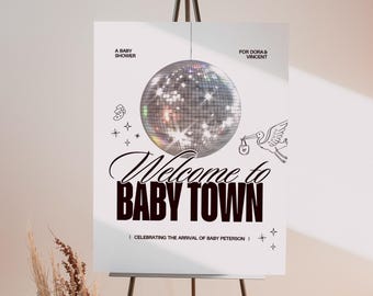 May include: A white baby shower sign on a black easel. The sign has a disco ball graphic and text that reads "Welcome to BABY TOWN" and "A BABY SHOWER FOR DORA & VINCENT". Decorative elements include stars and a stork.