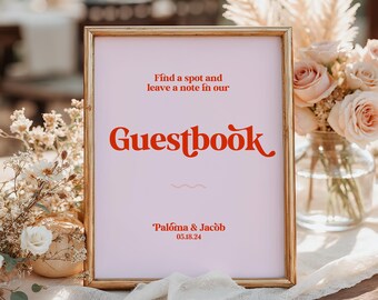 May include: A framed sign with the word "Guestbook" in red script. The sign has a light pink background and gold frame. The text "Find a spot and leave a note in our" is above the word "Guestbook". The names "Paloma & Jacob" and the date "05.18.24" are at the bottom.
