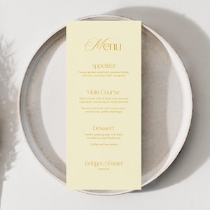 May include: A wedding menu card with gold lettering on a cream background, placed on a gray plate. The menu lists an appetizer, main course, and dessert. The names "Bridget & Daniel" and the date are at the bottom.