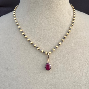 May include: A gold-toned necklace with a pendant featuring a ruby-colored oval gemstone. The necklace has a beaded design with gold-colored beads and a darker chain. The pendant is suspended from a delicate gold bail.