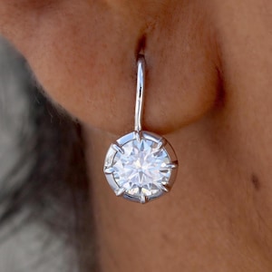 Puede incluir: Primer plano de un pendiente de plata con una gema grande, redonda y transparente. La gema está engastada en un bisel de plata con múltiples puntas. El pendiente cuelga de un gancho de plata curvado.