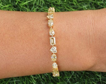 May include: A gold bracelet with various shaped diamonds, including round, pear, and emerald cuts. The bracelet is set on a gold chain and is displayed on a person's arm against a background of green grass.