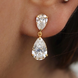 May include: A pair of gold drop earrings featuring pear-shaped, clear gemstones. The earrings have a small, pear-shaped stone at the top and a larger one dangling below, connected by a delicate gold chain. The gemstones are set in gold prongs.
