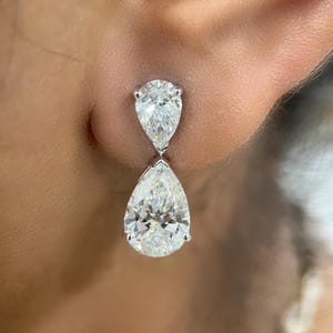 May include: A pair of elegant diamond drop earrings. Each earring features a pear-shaped diamond, with a smaller diamond above a larger one. The diamonds are set in a silver or white gold setting, reflecting light.