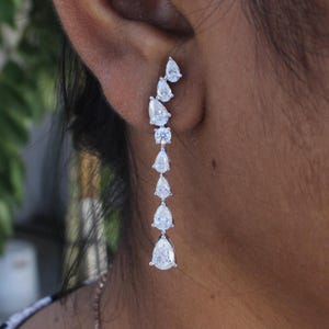 May include: A dangling earring featuring a series of clear, pear-shaped and round-cut cubic zirconia stones. The stones are set in a silver-colored metal, creating a cascading effect. The earring is worn on the ear.