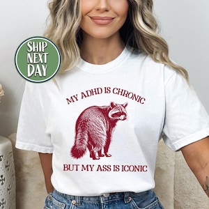 Puede incluir: Camiseta blanca con el texto "MY ADHD IS CHRONIC BUT MY ASS IS ICONIC" y un gráfico de mapache rojo. La camiseta es de estilo casual.