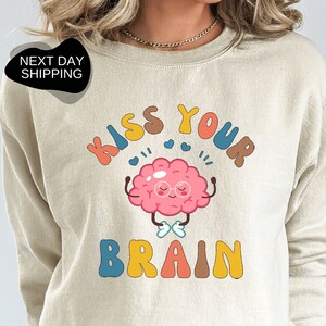 Może przedstawiać: Beżowa bluza z nadrukiem różowego mózgu z oczami i ramionami, z napisem "Kiss Your Brain" kolorowymi literami.