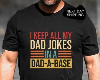 Op de afbeelding: Zwart T-shirt met de tekst "I KEEP ALL MY DAD JOKES IN A DAD-A-BASE" in retrostijl. Het shirt heeft een ronde hals en korte mouwen. Een golfkar is ook zichtbaar in de afbeelding.