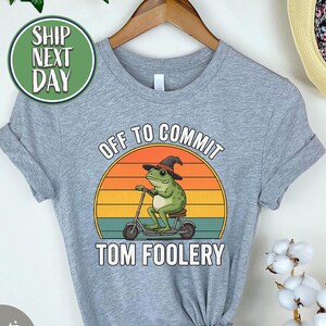 Könnte beinhalten: Graues Athletic Heather T-Shirt mit einem Frosch mit Hexenhut auf einem Roller, vor einem Sonnenuntergangsdesign. Der Text lautet "OFF TO COMMIT TOM FOOLERY". Das T-Shirt hängt an einem Holzkleiderbügel.