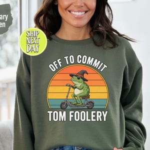 May include: Military green sweatshirt featuring the text "OFF TO COMMIT TOM FOOLERY" and a graphic of a frog on a scooter. The frog wears a witch's hat against a sunset. The sweatshirt has a crew neck.