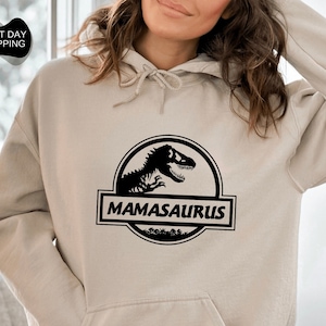 May include: A sand-colored hoodie with a black dinosaur graphic and the text "MAMASAURUS" printed on the front.