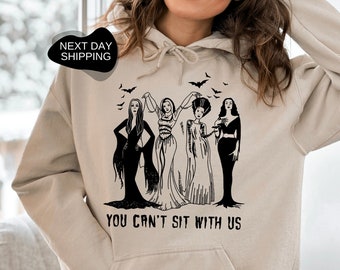 May include: A sand-colored hooded sweatshirt with a black graphic of four women in witch costumes. The text "You can't sit with us" is printed below the graphic.