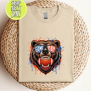 May include: A beige t-shirt with a bear graphic wearing sunglasses that read "GAME DAY" and holding a football. The design has red, white, and blue accents. The t-shirt is folded on a woven, circular surface.