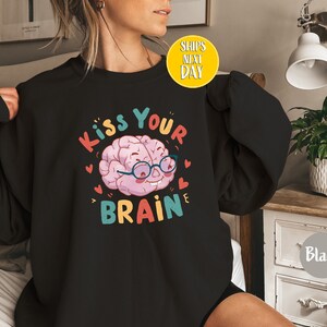 Może przedstawiać: Czarna bluza z kolorową grafiką mózgu w okularach i tekstem "Kiss Your Brain".