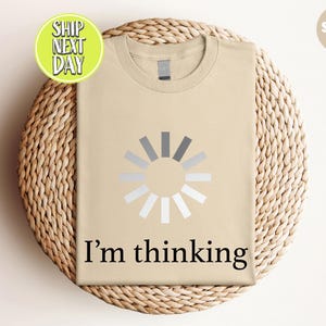 May include: A sand-colored t-shirt with a graphic of a loading symbol and the text "I'm thinking" in black. The shirt is displayed on a woven, circular surface.