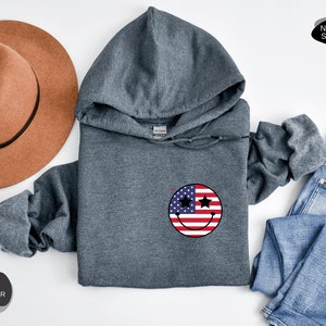 May include: Dark heather grey hooded sweatshirt with a smiley face design featuring the American flag. The smiley face has two stars for eyes.
