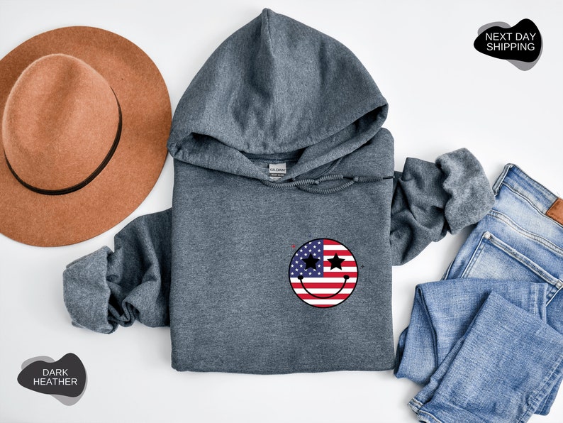 May include: Dark heather grey hooded sweatshirt with a smiley face design featuring the American flag. The smiley face has two stars for eyes.