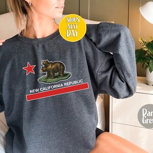 Peut inclure: Sweat-shirt gris fonc&eacute; avec un graphique du drapeau de l'&Eacute;tat de Californie repr&eacute;sentant deux ours et une &eacute;toile rouge. Le texte "NEW CALIFORNIA REPUBLIC" est imprim&eacute; sous le drapeau.