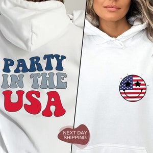 May include: White hoodie with a red, white, and blue "Party in the USA" graphic on the back and a smiley face with an American flag design on the front.