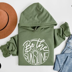 Pu&ograve; includere: Una felpa con cappuccio verde con il testo "Be the Sunshine" in un cerchio bianco sul davanti. La felpa &egrave; appoggiata su una superficie bianca con un cappello marrone e un paio di jeans blu.