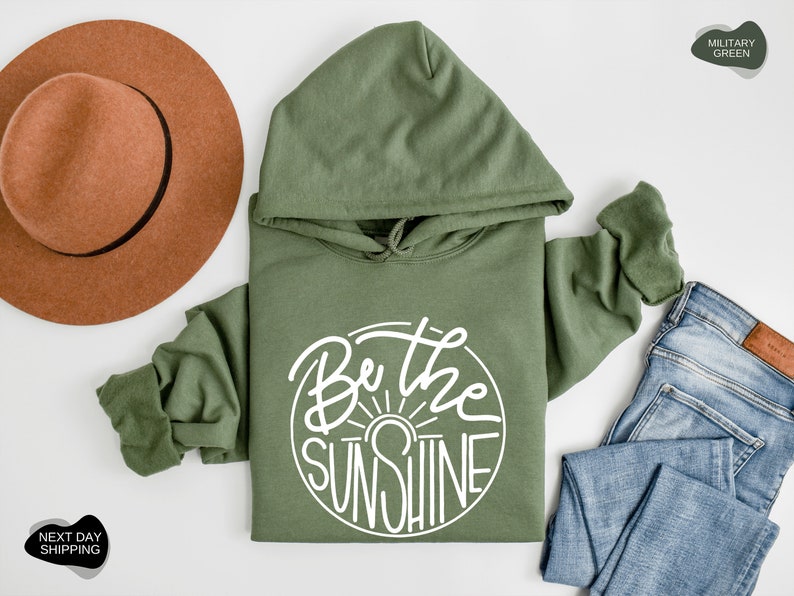 Pu&ograve; includere: Una felpa con cappuccio verde con il testo "Be the Sunshine" in un cerchio bianco sul davanti. La felpa &egrave; appoggiata su una superficie bianca con un cappello marrone e un paio di jeans blu.