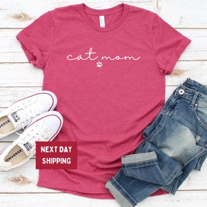 May include: Heather raspberry t-shirt with the text "cat mom" and a paw print. The shirt is paired with blue jeans and white sneakers. The image is taken on a light wood background.