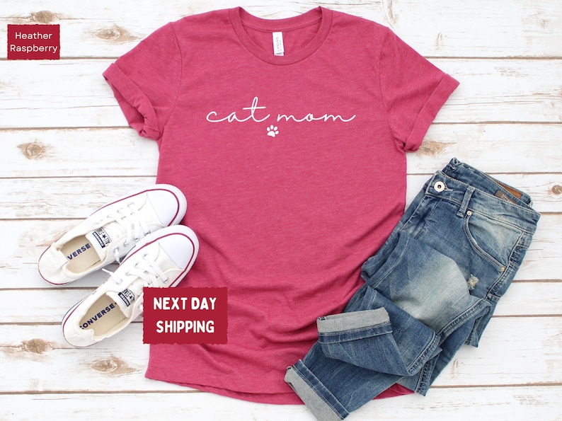 May include: Heather raspberry t-shirt with the text "cat mom" and a paw print. The shirt is paired with blue jeans and white sneakers. The image is taken on a light wood background.
