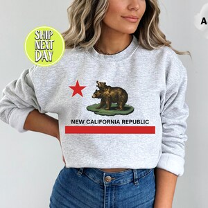 Peut inclure: Un sweat-shirt gris avec une &eacute;toile rouge et deux ours bruns sur une colline verte. Le texte "NEW CALIFORNIA REPUBLIC" est imprim&eacute; sous les ours.