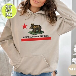 Peut inclure: Un sweat &agrave; capuche couleur sable avec une &eacute;toile rouge et deux ours bruns sur le devant. Le texte "NEW CALIFORNIA REPUBLIC" est imprim&eacute; sous les ours.