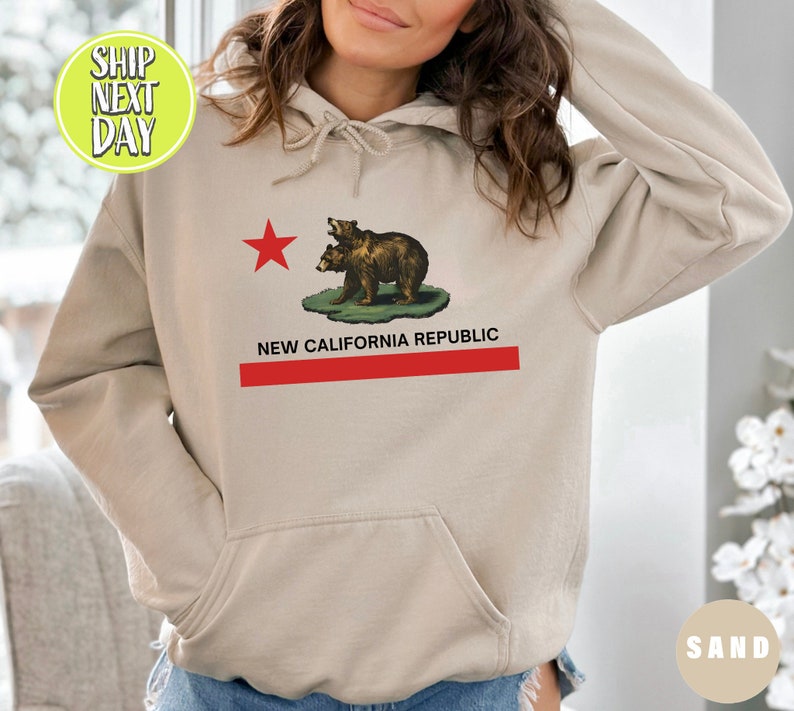 Peut inclure: Un sweat &agrave; capuche couleur sable avec une &eacute;toile rouge et deux ours bruns sur le devant. Le texte "NEW CALIFORNIA REPUBLIC" est imprim&eacute; sous les ours.